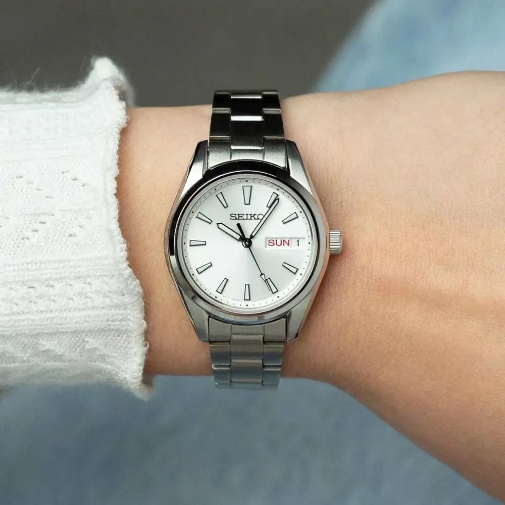 Muñeca de mujer con un reloj SEIKO clásico de acero, esfera plateada con día y fecha, junto a un suéter blanco